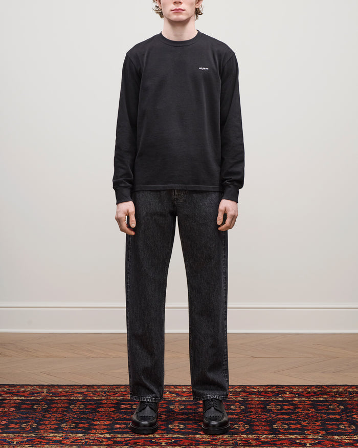 A person stands on a patterned rug wearing Relaxed Fit 5-Pocket Denim Pant, black sweatshirt, and black shoes.