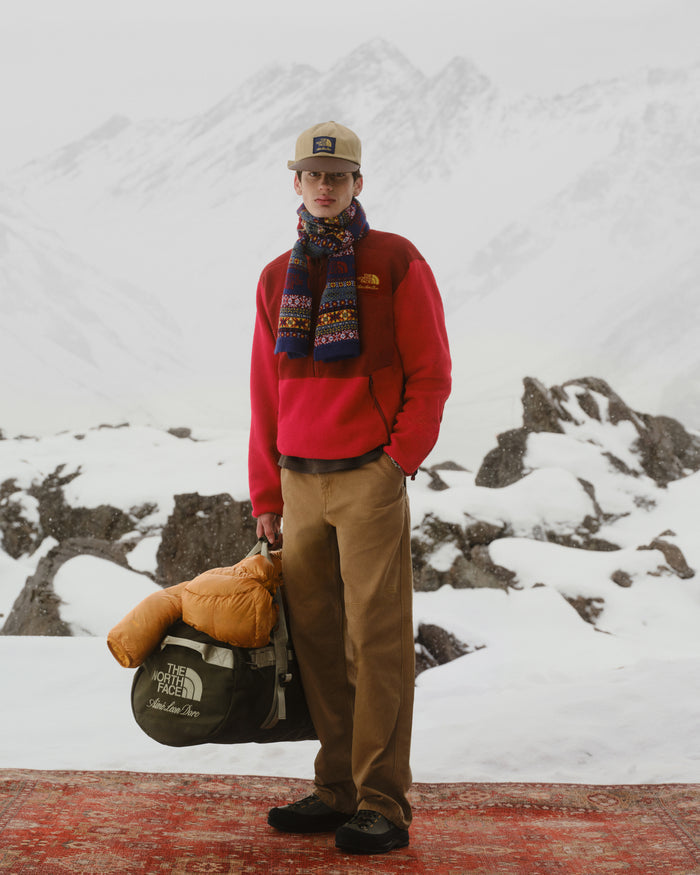 Man in red jacket and ALD/The North Face Fair Isle Scarf stands on snowy mountain, holding a duffel and sleeping bag.