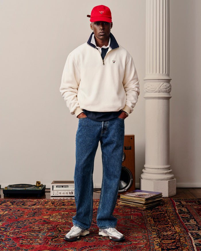 A man in a white pullover, blue jeans, and Crest Logo Hat stands on a patterned rug by speakers and a record player.