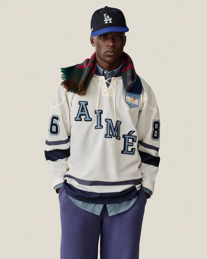 Man in Stone Washed Marina Sweatpants, LA cap, plaid scarf, AIMÉ 86 white hockey jersey, hands in pockets.