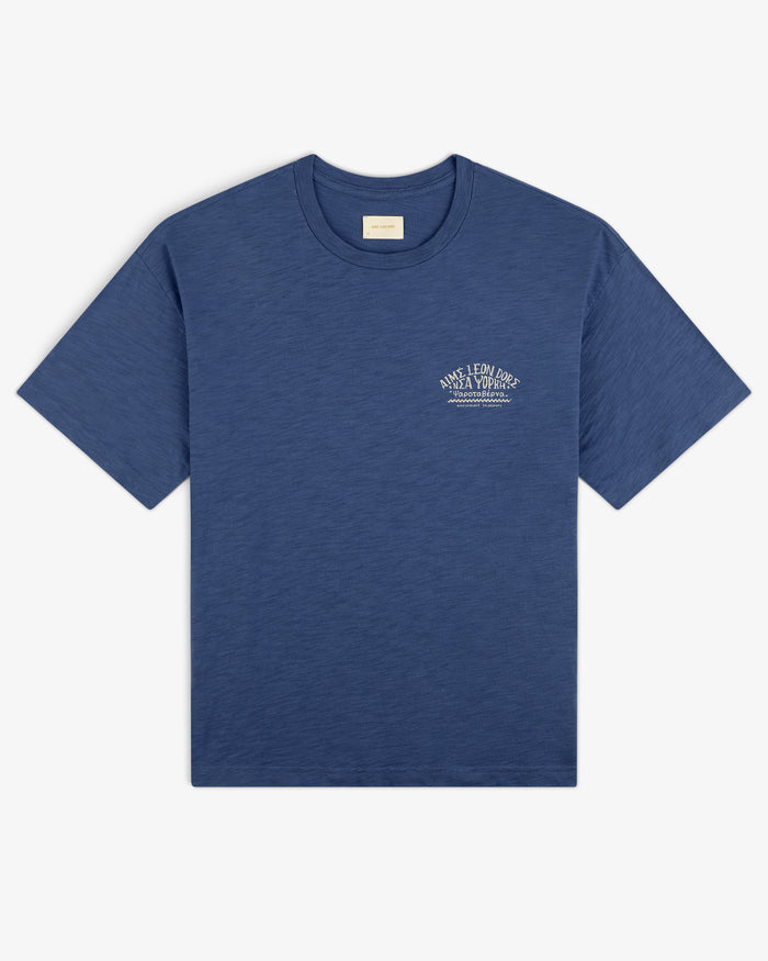 Taverna Tee: Blue short-sleeve t-shirt with small white text graphic on left chest, shown on white background.