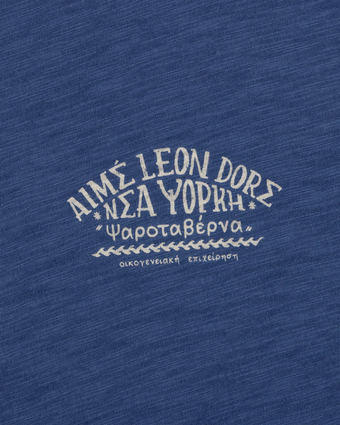 White Greek text and AIMÉ LEON DORE on blue textured fabric, featured on the Taverna Tee.