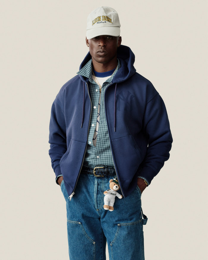 Man in Yankees Washed Full-Zip Hoodie, checkered shirt, cap, loose jeans, and teddy bear keychain on his belt.